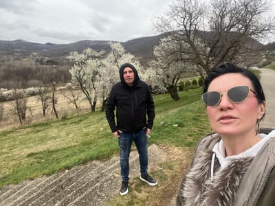 Two individuals, a woman and a man, are positioned outdoors on a grassy slope next to concrete steps under an overcast sky, in what appears to be Despotovac Municipality, Serbia. The woman, on the right in the foreground, has dark hair and wears dark, gold-framed cat-eye sunglasses. She is wearing a white hooded sweatshirt beneath an olive-green jacket with a prominent grey faux fur collar and is taking the photograph, indicated by her arm position and the framing. Her cheeks show visible pink blush, and she wears gold-colored drop earrings. The man, positioned slightly behind and to the left of the woman, wears a black hooded jacket with the hood up, distressed blue jeans with white splatters, and black athletic shoes with white soles. He stands with his hands in his pockets, holding a lit cigarette in his right hand. Both individuals look directly towards the camera.
The background features rolling hills covered predominantly with bare deciduous trees, interspersed with trees displaying white blossoms. A valley with scattered structures and a brown field is visible between the foreground and the distant hills. A paved road curves through the right background, with several parked vehicles visible. A small, dark-roofed structure is partially visible amidst the blossoming trees behind the man. The immediate foreground includes concrete steps on the left and a sloped area of green and tan grass.