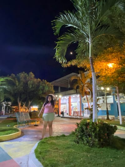 The image displays a nighttime scene within a public plaza in General Villamil Playas, Ecuador. A female individual stands in the foreground, slightly left of center, positioned on a multi-colored paved pathway with sections of yellow, blue, pink, and gray. She has dark, wavy hair and wears a light pink short-sleeved top and light green shorts; her right hand is raised near her head.
The background features a two-story building appearing commercial, brightly illuminated with internal white lights and exterior accents. Its ground floor exhibits a white facade with vertical orange elements, large display windows showing merchandise, and two red slatted metal benches positioned outside. To the far right, a blue building with white trim is partially visible, displaying the text "PLA."
The environment includes numerous trees, with a prominent palm tree in the mid-right foreground and additional palm trees and deciduous trees with green foliage throughout. Artificial lighting, primarily from streetlights, casts a warm yellow-orange glow on much of the foliage and the ground. Patches of green grass and dense shrubbery are present in the foreground and mid-ground, including a specific shrub with small red accents in the lower right. A light blue slatted metal bench is located in the mid-ground to the left of the central figure. A crescent moon is visible in the dark night sky in the upper center-right. Small, indistinct figures are discernible within the illuminated background building.