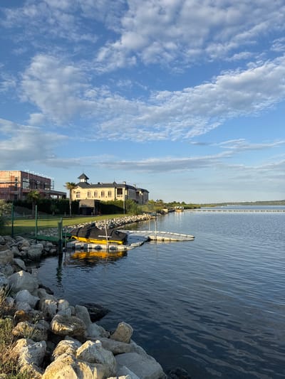 The image captures a serene waterside view in Kazashko, Bulgaria, under a bright sky dotted with white, fluffy clouds. The foreground is dominated by a rocky embankment, with large, light-colored stones and some sparse vegetation. Adjacent to the embankment is a floating dock with a yellow jet ski covered by a black tarp.
To the left, a green fence separates the embankment from a manicured lawn. Beyond the lawn, two buildings are visible. The building on the left appears to be under construction, with scaffolding and exposed brickwork. To its right stands a larger, more elegant structure with a light-colored facade, multiple windows, and a dark, conical roof topping a central tower. Palm trees and lush greenery surround the buildings, creating a sense of a well-developed waterfront property.
The water itself is calm, with gentle ripples reflecting the sky. In the distance, the expansive lake stretches towards the horizon, where a long, thin structure, possibly a pier or a breakwater, is visible. The overall atmosphere is tranquil, suggesting a peaceful day in this Bulgarian lakeside community.