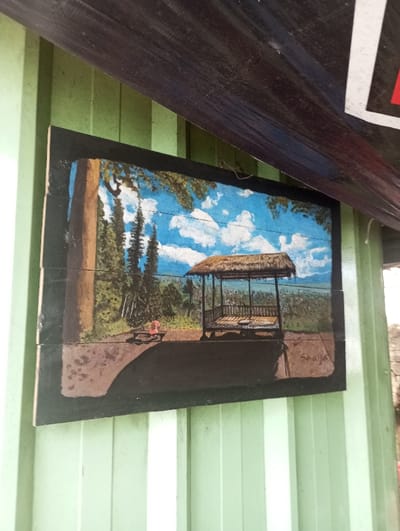 The image depicts a landscape painting mounted on a light-green, vertically corrugated wall in Gadog, Indonesia. The painting, rendered on what appears to be horizontal wooden panels, portrays a rural scene under a blue sky with white clouds. On the right side of the painting, a traditional Indonesian-style gazebo with a thatched roof stands on an elevated platform. To the left, a forest of tall, slender trees is visible, with a small red object (possibly a bench or small figure) positioned near the foreground. The background extends into a wide vista of lower-lying vegetation and distant hills. A signature, "Saaya" or "Swaya," is visible in the bottom right corner of the painting. A dark, possibly black, banner or canopy hangs diagonally across the upper portion of the frame, partially obscuring the top left of the painting and the wall above it. The banner exhibits a white rectangular element on its right side. No individuals are present in the scene.