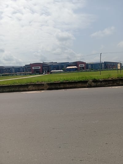 The image shows a wide asphalt road in Igwuruta, Nigeria, with a low concrete barrier separating it from a green lawn. In the background, a series of large industrial or commercial buildings are visible. The buildings are predominantly dark gray with some sections painted red. Utility poles and power lines run along the foreground of the buildings. The sky is partly cloudy with a mix of white and gray clouds. A signpost stands on the far right of the image. No people are clearly visible.