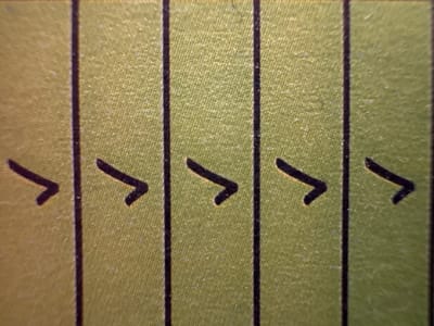 The image shows a close-up view of a surface with vertical black lines, creating distinct sections. Within these sections, a pattern of dark, arrow-like shapes is visible, pointing to the right. These shapes appear to be hand-drawn or printed, with a slightly rough texture. The background surface is a textured, greenish-yellow color, reminiscent of astroturf or a woven material. The lighting is even, with no strong shadows, suggesting an indoor or overcast environment. There is no discernible text in the image. The overall impression is of a diagram or a visual aid, possibly related to a game, sport, or instructional material. The specific context of Bydlino, Poland, does not provide direct visual cues within the image itself, but suggests a possible geographical setting for the origin or use of this object.