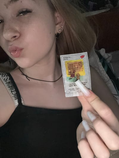 This is a close-up, vertically oriented, top-down photograph of a young person's face and hand. The person has blonde hair, light skin, and is wearing makeup, including noticeable eyelashes and lip gloss. They are also wearing a black choker with rhinestones and a necklace. The person is holding a small, rectangular card with text and an illustration. The illustration depicts two cartoonish characters, possibly from a children's story, within a yellow-bordered frame. A red heart is visible in the upper left corner of the card. Text on the card appears to be in Russian and English, with phrases like "Любовь это..." and "Love is...". The background is dark and out of focus, suggesting an indoor setting. The lighting is dim. The overall mood is intimate and personal. The provided location context is Velykyi Sukhodil, Ukraine.