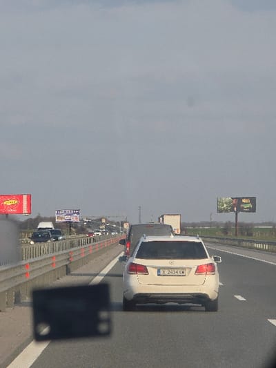The image depicts a multi-lane highway scene in Opalchenets, Bulgaria, under an overcast sky. The viewpoint is from inside a vehicle, indicated by a dark, out-of-focus rectangular object with two circular elements in the lower-left foreground. The asphalt road features white solid and dashed lane markings. A metal guardrail, white with orange vertical reflectors, lines the left shoulder. A queue of vehicles is present, positioned in the same direction as the viewer. The primary vehicle in view is a white Mercedes-Benz station wagon, license plate X 2434 KW (Bulgaria), positioned in the rightmost visible lane. Immediately ahead of the Mercedes are a dark grey van and a white semi-truck trailer. Additional vehicles are visible in the left lanes and beyond the guardrail. Billboards are present along the road: a red one with yellow elements on the far left, another displaying "TOP ЦЕНА!" advertising, and a large dark-themed advertisement on the right. The background consists of flat, vegetated terrain and a distant treeline. An overhead highway structure is visible in the far distance. No human figures are clearly identifiable within the frame.