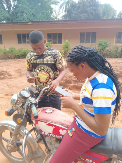 Two young adults are engrossed in their phones while standing beside a weathered red motorcycle. The man is wearing a black and gold patterned shirt, and the woman is seated on the motorcycle with a blue and white striped shirt and burgundy pants. The bike's brand, "DAYLONG," is visible on the gas tank. A building with windows stands in the background, and foliage is sparsely scattered around the scene. The overall setting appears to be outdoors.