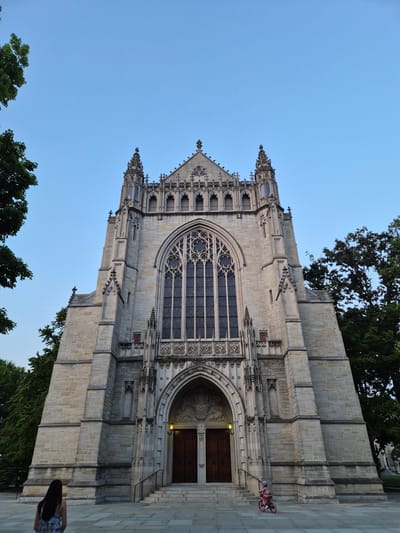 The image depicts the exterior of a large, gothic-style stone building, likely a chapel or cathedral, under a clear, pale blue sky. The architecture features intricate stonework, including pointed arches, detailed carvings, and ornate spires. A large, elaborate stained-glass window dominates the central facade above the main entrance. The entrance itself is framed by a large arched doorway with heavy wooden doors.

In the foreground, to the left, a person with long dark hair, wearing a patterned dress, is walking away from the camera towards the building. To the right, a small child wearing a hat and riding a pink bicycle stands on the paved area in front of the building. The building is flanked by mature green trees. The ground in front of the building is paved with large, grey flagstones. The building appears to have decorative stonework with the inscription "1925" visible on the lower right section.

This building is Princeton University's Graduate College, located in Princeton, New Jersey, United States of America.