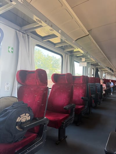 This is a photograph taken inside a train carriage, looking forward along the aisle. The seats are upholstered in a deep red velvet fabric with a subtle pattern. The carriage appears to be mostly empty, with a few passengers visible in the distance. The lighting is bright, suggesting it is daytime. The overall impression is one of a well-maintained, albeit perhaps older, train interior. The location in the city of Cherna Gora, Bulgaria, is not discernible from the image itself.