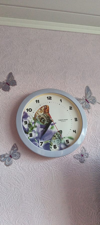 An indoor shot shows a round wall clock with a light gray frame and a white face. The clock has black Arabic numerals indicating the hours, with smaller tick marks for minutes. The clock face features an image of two butterflies and purple flowers. The hour and minute hands are black, and there is a thin red second hand. The clock is surrounded by four decorative, translucent butterflies with glitter accents attached to a light pink textured wallpaper. The wallpaper has a raised, swirling pattern. A white ceiling molding is visible at the top of the image.