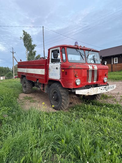 The image depicts a red fire truck with white stripes parked on a dirt path in a grassy area. The fire truck is the primary subject, positioned centrally and facing slightly to the right. It has large, rugged tires, a typical fire truck cab with visible windshield wipers and a blue flashing light on top. Text is visible on the side of the truck, appearing to be in Cyrillic script. The license plate on the front bumper is also visible and readable.
In the background, there is a wooden house with a dark roof to the right of the fire truck. To the left and behind the truck, a tall, slender tree with light-colored bark is prominent, with other trees and foliage forming a dense green backdrop. Electrical poles and wires are visible in the sky, suggesting a rural or semi-rural setting. The ground is a mix of dirt and tall green grass, with some yellow wildflowers growing in the foreground. The sky is overcast, with cloudy conditions. The scene appears to be outdoors, in what is likely a village or countryside setting. No people are present in the image. The overall impression is one of a still, quiet moment, possibly in a village like Кукуи, Russia, given the context of the provided information.