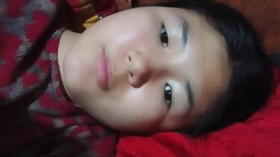 This media file is a close-up, overhead shot of a young person lying down. The person has dark hair, dark eyes, and light skin with some visible blemishes. They are wearing a red and grey patterned garment. The background is out of focus, but appears to be a red surface, possibly a blanket or pillow, and a darker, wooden surface on the right. The lighting is dim, suggesting it might be indoors during the evening or night. The person's expression is neutral, with their eyes open and looking towards the camera. The context of Uchkun, Kyrgyzstan, indicates a rural or mountainous setting. There is no visible text in the image.