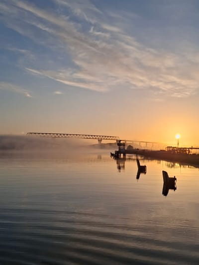 The image depicts a sunrise or sunset over the Kiel Canal in Brunsbüttel, Germany. The sky transitions from blue with streaky clouds to a warm orange hue near the horizon where the sun is visible. Industrial structures, including cranes and other port facilities, are silhouetted against the bright sky. The water of the canal reflects the colors of the sky and the shapes of the industrial elements. There are no people visible in the image.