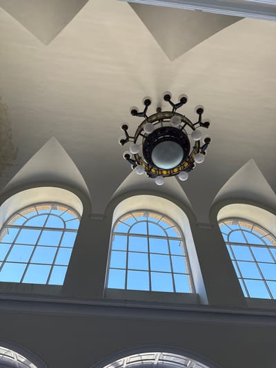 The image presents an upward perspective of a grand interior ceiling, focusing on a large, elaborate chandelier and several arched windows. A decorative chandelier with a central spherical light and numerous smaller, white globe-shaped bulbs arranged in a circular pattern hangs prominently from the ceiling. The chandelier's frame features intricate detailing with what appears to be colorful stained glass or patterned elements.
The ceiling, painted in a light, neutral tone (likely off-white or light beige), showcases architectural elements like subtle vaults and angular sections. Along the right side of the frame, three large, arched windows are visible, allowing abundant natural light to flood the space and revealing a clear, vibrant blue sky outside. These windows are divided into multiple rectangular panes by a grid of white mullions. The windows are set within deep, curved recesses in the ceiling structure, creating distinct patterns of light and shadow on the surrounding surfaces. Brickwork is faintly visible around the exterior frames of some windows.
The photograph captures a sense of architectural grandeur and the natural illumination of the space during clear daytime weather. No people or specific activities are depicted, and no text is visible within the frame. The scene suggests a public building or a large, ornate hall, consistent with a location in Szeged, Hungary.
