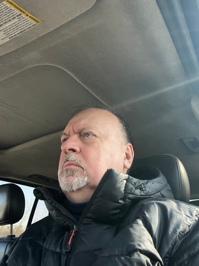 An adult male, appearing middle-aged or older with a fair complexion, balding scalp, and a grey goatee, is depicted seated inside a vehicle in Ерино, Russia. He wears a black puffy winter jacket over a dark turtleneck and is looking upward and to his left. The vehicle's interior features a light grey headliner, and a yellow warning label ("AVERTISSEMENT / WARNING") is partially visible on the sun visor. Strong sunlight illuminates the scene from the right. A dark headrest is behind the subject. The exterior visible through the window on the left shows bare tree branches against a bright sky.