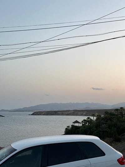 In Bolnuevo, Spain, a white car is parked on a hillside overlooking the sea. The car's rear is in the foreground, with its roof and rear window visible. Beyond the car, the calm sea stretches out, meeting a distant, hazy coastline with rolling hills. A small gazebo-like structure is nestled amongst lush green trees and palm trees on the slopes leading down to the water. Power lines are strung across the top of the image, silhouetted against a pale, twilight sky. The overall scene suggests a peaceful, coastal setting at dusk.