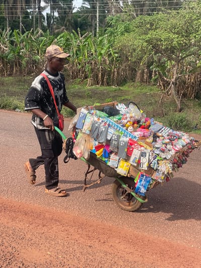 A man walks along a paved road in Ogbe, Nigeria, pulling a heavily laden, single-wheeled cart. The cart is filled with a variety of goods, including phone cases, locks, and plastic containers. The man is wearing a patterned shirt, black pants, sandals, and a hat. He is pulling the cart with a green handle. The road is surrounded by vegetation.