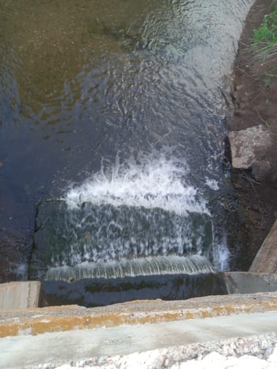 A top-down shot captures a small waterfall cascading over a concrete structure. The water is frothy and white where it falls, creating a dynamic contrast with the darker, calmer water below. The surrounding area appears to be a natural setting, with a dirt bank covered in sparse green vegetation visible on the left. Some rocks are present along the edge of the bank, and a larger, rough-hewn stone sits near the water's edge. On the right side of the image, a rough, textured concrete wall with a yellowish-brown stain runs vertically, suggesting it's part of an embankment or dam. The scene is likely outdoors, given the natural elements and the flow of water. The lighting suggests it could be daytime, but no specific time or weather conditions are evident. There is no visible text in the image.