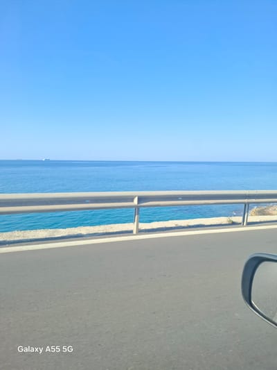 A partially rotated eye-level shot captures a road alongside the sea, likely taken from a moving vehicle, as evidenced by the presence of a side mirror. The road occupies the right side of the frame, with white lines indicating lane markings. Adjacent to the road is a walled pathway, separating the road from a narrow strip of beach. The sea extends to the left of the pathway, meeting the horizon under a clear blue sky. The lower right corner of the image includes the text "Galaxy A55 5G," indicating the device used for the photo. The location is plausibly near Gouves Municipal Unit, Greece, given the coastal characteristics and the context.