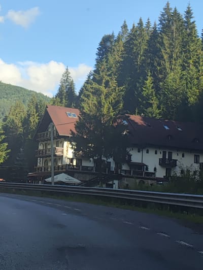 In Borșa, Romania, a three-story hotel with a dark red, gabled roof is nestled amongst dense evergreen trees. The hotel's facade is white, featuring numerous balconies with wooden railings and small, dark-paned windows. Several skylights are visible on the roof. A light-colored umbrella is set up outside the hotel on the ground level, near what appears to be an outdoor seating area. The hotel is situated on a slope, with the foreboding forest dominating the background. In the foreground, a wet asphalt road runs parallel to the hotel, separated by a metal guardrail and a strip of green grass. The sky is a clear blue with scattered white clouds.