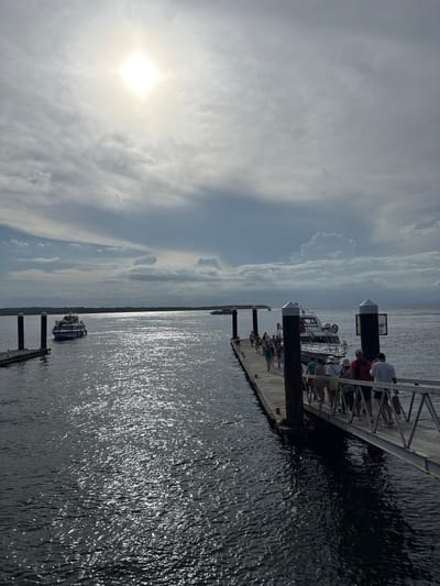 A landscape oriented photograph shows a dock jutting into the water on a partially cloudy day. The sun is high in the sky and reflecting on the water. There are people standing and sitting on the dock near several boats. A third boat is closer to the bottom of the image and further away from the dock. The photograph was taken in the city of Toyapakeh, Indonesia.