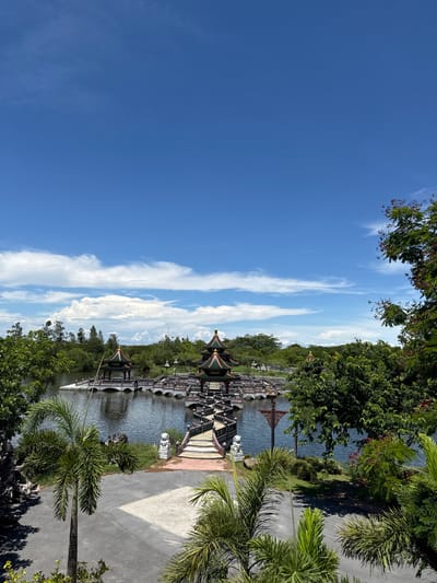 The image depicts a scenic view in Bang Pu Subdistrict Municipality, Thailand. The right side features lush vegetation with various trees, including palm trees, bordering a paved pathway. To the left of the pathway is a body of water, on which a series of intricately designed covered bridges are constructed, connecting several ornate structures. The bridges and buildings exhibit a distinct architectural style, possibly of Chinese or Thai influence, with detailed roofs. The sky, dominating the left portion of the image, is a clear blue with wispy white clouds. No people are visible in the frame.