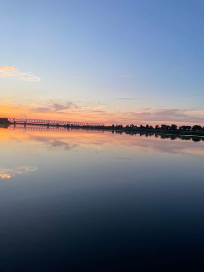 A wide, calm river reflects the sky and a distant railway bridge at sunset. The sky transitions from a soft blue at the top to warm hues of orange and pink near the horizon, with scattered wispy clouds. A long, truss railway bridge stretches across the river, its structure clearly visible and mirrored in the water below. The far bank of the river is lined with a dense row of trees, forming a dark silhouette against the colorful sky. The water is dark and smooth, creating a near-perfect reflection of the scene above, with subtle ripples disturbing the otherwise still surface. The overall impression is one of serene natural beauty at dusk. This scene is likely the river Neman in Sovetsk, Russia, given the prominent railway bridge.