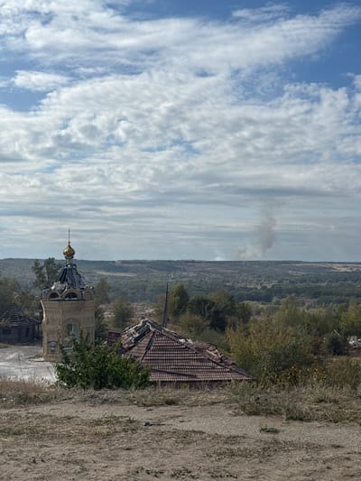 This image captures a panoramic view from an elevated position in Izium, Ukraine. The foreground consists of dry, uneven terrain with sparse light-brown and green vegetation. In the midground, several structures are visible: a prominent, ornate light-colored brick bell tower or chapel, topped with a partially damaged golden dome and cross, revealing a blue-tiled surface beneath. To the right of this structure, a building with a reddish-brown tiled roof shows significant damage, with numerous tiles missing and portions of the roof structure exposed. To the left of the bell tower, a smaller, heavily damaged wooden structure with an exposed, dilapidated roof frame is present. Green and yellowish-brown trees and bushes are interspersed around these buildings and extend into the background. The background features a vast expanse of forested hills and valleys stretching to the horizon. In the distant background, a distinct column of light gray smoke rises vertically before dispersing horizontally. The sky above is partially overcast with numerous white and gray clouds. No individuals are present or discernable in the scene.