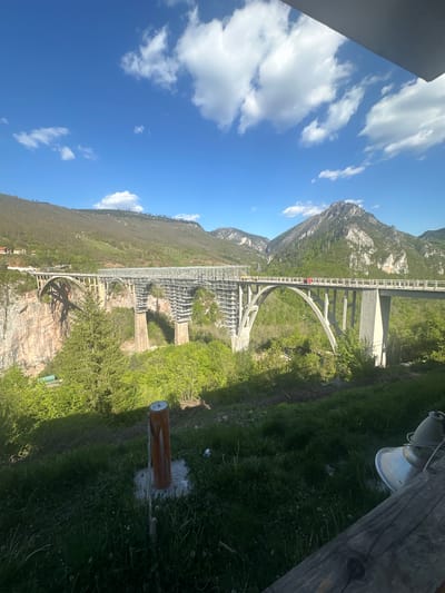 The image captures a view of the Đurđevića Tara Bridge in Montenegro. The bridge spans a deep canyon with the Tara River flowing at the bottom. The mountainous terrain is visible in the background, with a clear blue sky dotted with white clouds overhead. The bridge is a concrete arch structure with multiple spans. Vegetation covers the canyon walls and surrounding landscape. A lamp and a wooden post are visible in the foreground, suggesting the viewpoint is from a structure adjacent to the bridge. The scene appears to be taken during daylight hours.