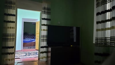The image shows an interior view of a room in Yakkabog, Uzbekistan. A doorway on the left leads into another room with a bed and a wooden door. A large black television is mounted on a dark cabinet in the foreground. The room is decorated with striped curtains on the left and right sides. The walls are painted green. A rug with a pattern is visible on the floor.
