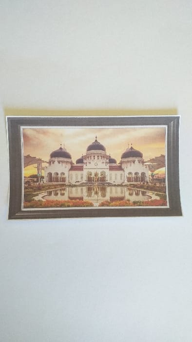 The image displays a rectangular color print, ostensibly a photograph, placed horizontally on a uniformly light-colored, flat surface. The print depicts a grand mosque, identified as the Baiturrahman Grand Mosque, presented as a scene from Limpok, Indonesia.

The mosque features a prominent central dome flanked by two smaller domes, all dark in color, surmounting white, multi-arched structures. Its ornate facade is reflected clearly in a still body of water that occupies the foreground. Surrounding the reflective pool are landscaped areas with green foliage and patches of reddish-orange flora, suggesting flowerbeds. The sky above the mosque exhibits warm, diffused light with orange and yellow gradients, indicative of either sunrise or sunset. Large, white, inverted-V or umbrella-like structures are visible to the left and right of the main mosque building, positioned over what appear to be paved courtyards or walkways. No human figures are present within the depicted scene.

The print itself includes a simulated dark frame with a thin white internal border. The right edge of the print exhibits a slight upward curl.