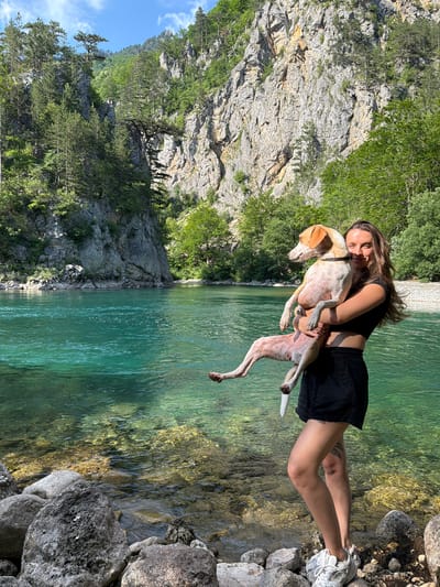 A young woman with long, brown hair, wearing a black crop top and black shorts, holds a medium-sized, light-colored dog with tan markings in her arms. The woman is smiling at the camera. The dog is looking to its left. They are standing on the rocky shore of a crystal-clear turquoise river. In the background, a steep, rocky mountain covered in dense green trees rises from the opposite bank of the river. The sky above is blue with scattered white clouds. The scene depicts a natural landscape likely in or near the city of Bistrica, Montenegro, known for its mountainous terrain and rivers.