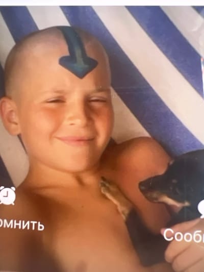 This is a close-up, horizontally oriented photograph featuring a young boy with a shaved head. A blue arrow is painted on his forehead, pointing towards the right side of the frame. He is looking slightly upwards and to his right, with a gentle smile on his face, suggesting a pleasant or playful mood. The boy appears to be wearing a blue and white striped garment or lying on a striped surface.
Partially visible in the upper right corner is a small black dog, likely a dachshund, being held by someone whose hand and arm are also visible. The hand is positioned around the dog's back, and the dog is looking towards the boy.
In the bottom right corner, there is a circular icon resembling an alarm clock, with the text "помнить" (which translates to "remember" in Russian) written vertically to its right. In the upper right corner, the word "Сообщ" (likely an abbreviation for "сообщение," meaning "message" in Russian) is also visible.
The lighting suggests it is daytime, and the overall atmosphere is casual and intimate, possibly indicating a personal moment captured at home or during a relaxed outing. The location context of Bernex, Switzerland, does not provide any further specific visual cues within the image itself.