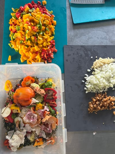 The scene depicts a food preparation area on a grey countertop surface in Anderlecht, Belgium. In the upper left quadrant, a teal cutting board is laden with a substantial pile of finely diced bell peppers in shades of red, yellow, and green. A single, small yellow pepper piece rests detached from the main pile on the board. In the lower left, a transparent plastic container is filled with various vegetable trimmings and organic waste, including bell pepper cores and seeds, onion peels, garlic skins, and other discarded vegetable pieces, indicating active food preparation. A partially intact orange bell pepper and several green and red pepper remnants are visible within the scraps. In the right half of the image, a dark grey cutting board (bearing faint "IKEA" branding) holds three distinct piles of diced ingredients: a top pile of minced garlic, a middle pile of diced white onions, and a bottom pile of diced light brown ingredients. In the upper right, a second, smaller teal cutting board is visible, featuring a silver-bladed kitchen knife resting horizontally with its handle oriented to the right. No individuals are present in the image.