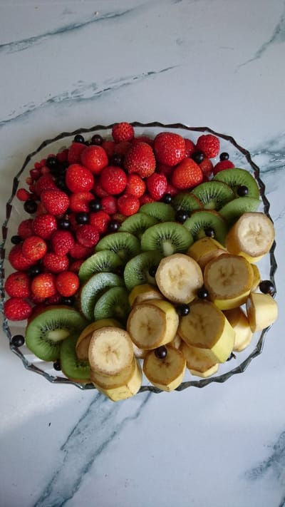 A clear glass platter, with a decorative, rippled edge, is filled with a colorful assortment of fruits, placed on a light-colored marble countertop. The platter is arranged with three distinct sections. On the left side, numerous small, red strawberries, some whole and some halved, are interspersed with dark, small berries, likely currants or blueberries. In the middle section, several circular slices of kiwi fruit are arranged in a slightly overlapping fan pattern, revealing their characteristic green flesh with dark seeds. To the right, slices of banana, cut into rounds, are stacked and fanned out. Some of the banana slices show a slightly browned edge. Small, dark berries are also scattered among the banana slices. The overall scene is well-lit, highlighting the fresh appearance of the fruit. There are no people visible in the image, and the setting does not provide any specific information that would suggest a location in Dvirets, Ukraine.