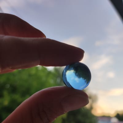 A close-up shot focuses on a person's thumb and index finger holding a translucent blue marble. The marble reflects the sky and surrounding greenery, creating a miniature world within its surface. The background suggests an outdoor setting, with blurred green foliage and a hazy sky indicating it might be a partly cloudy day. The lighting on the fingers and the marble suggests it is either late afternoon or early morning, possibly during sunset or sunrise, given the warm tones in the sky. The person's fingers are silhouetted against the lighter background. There is no visible text in the image. The setting is Kineshma, Russia.