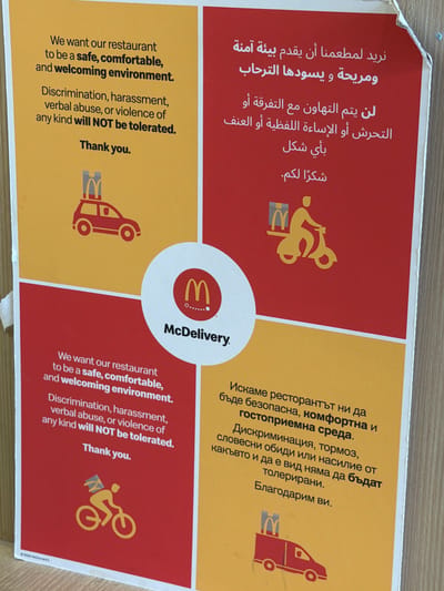 The image is a sign from McDonald's with a message about creating a safe and welcoming environment. The message is translated into three languages: English, Arabic, and Bulgarian. The sign states that discrimination, harassment, verbal abuse, or violence of any kind will not be tolerated. Each section has a McDonald's logo or icon and is separated by a circle in the center with the words McDelivery. The sign is located somewhere inside the restaurant or drive-thru.