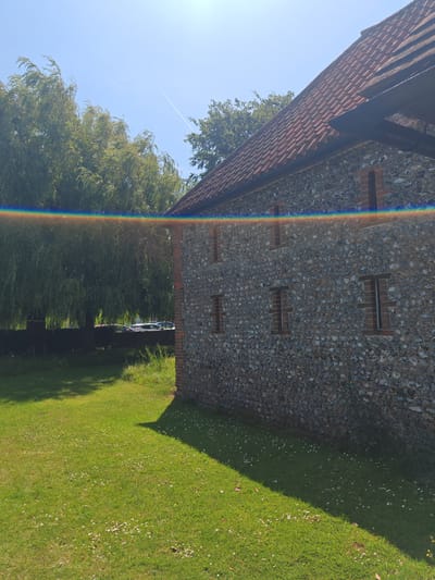 This image captures a sunny outdoor scene, likely in Worthing, United Kingdom, given the context. The foreground is dominated by a lush green lawn, scattered with small white flowers and dappled with shadows cast by the sun. A portion of a stone building, possibly an old barn or outbuilding, is visible on the right side of the frame. The building features a tiled, reddish-brown roof and walls constructed from grey stones interspersed with brickwork. Several small, rectangular windows are set into the stone facade. To the left of the building, a large weeping willow tree with dense, drooping branches stands prominently, its leaves a vibrant green. Beyond the tree, a row of parked cars is partially visible, suggesting a nearby parking area or road. The sky is a clear, bright blue, with a faint contrail from an aircraft visible high up. A rainbow-like lens flare streaks across the upper portion of the image, indicating the bright sunlight and the angle of the camera. The overall impression is one of a peaceful, sunlit day in a rural or semi-rural setting.