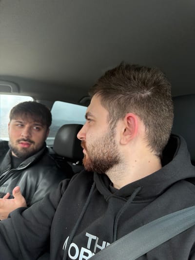 Two adult males are observed seated inside a vehicle. The scene is located in Gabarnitsa, Bulgaria.

The male in the foreground, positioned to the right, is seen in profile, facing left. He has short, dark brown hair and a reddish-brown beard. He is wearing a black hooded sweatshirt with a partial "THE NORTH FACE" logo in white visible on the chest. A grey seatbelt diagonally crosses his torso from his right shoulder to his left hip. His right ear exhibits a reddish hue. His gaze is directed towards the male in the background.

The second male, situated in the background to the left, is partially visible from the chest up, facing towards the right foreground male. He has dark, medium-length hair with bangs and a dark beard and mustache. He is dressed in a black, possibly leather or synthetic, puffer jacket. His right hand is raised, palm slightly upwards, indicating an active gesture, possibly during speech. His mouth is slightly open, suggesting he is speaking.

The interior of the vehicle features a dark grey headliner and black seat upholstery. The exterior environment visible through the windows is blurred, suggesting movement or a shallow depth of field, with an overcast sky.