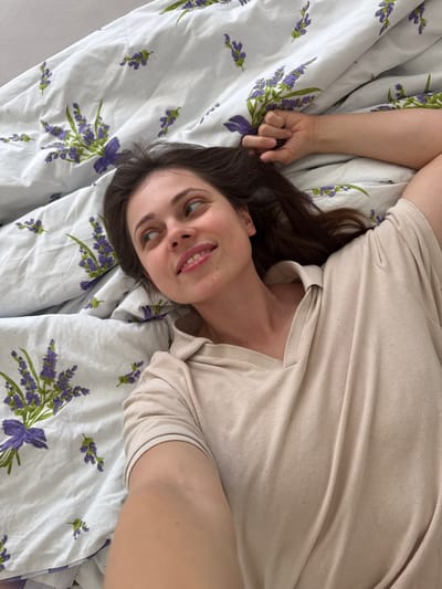 A woman lies on a bed with a white sheet patterned with sprigs of purple lavender. She is facing the camera, smiling slightly, and her dark hair is spread on the pillow. She wears a light beige, long-sleeved shirt. Her left hand is near her hair. The location in the city of Kőröshegy, Hungary is undetermined from the image.