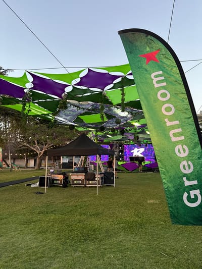 An outdoor event venue in Noumea, France, is depicted under a clear, bright sky, likely in the late afternoon or early evening. The scene features a large overhead canopy composed of interconnected green and purple fabric shade sails, from which artificial foliage and small mirrored spheres are suspended.
In the foreground, a prominent green feather banner stands upright on the right, displaying a red five-pointed star (Heineken logo) and the white text "Greenroom" oriented vertically. To the left of the banner, a black pop-up tent shelters an array of professional sound and lighting equipment, including multiple large cases labeled "1PLAY" and various consoles. A black mat pathway extends from the tent to the left across the green grass.
Several individuals are visible in the midground, near a large stage structure positioned towards the rear of the grassy area. These individuals appear to be engaged in setup or technical checks. The stage features a prominent LED screen displaying a stylized white letter "R" on a purple and green geometric background. Additional lighting rigs and decorative elements are integrated into the stage design. In the background, beyond the stage, trees and string lights are visible, with other distant structures suggesting a park or resort setting. The entire area is covered in well-maintained green grass.