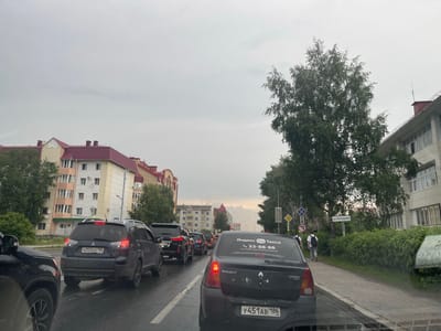 The image captures a traffic jam on a street in Khanty-Mansiysk, Russia, likely during daylight hours given the overcast sky. The wet asphalt suggests recent rain. In the foreground, the rear of a black car is visible, with a license plate reading "Y451AB 186". A decal on its rear windshield advertises "Yandex Go Taxi" with a phone number. The car's taillights are illuminated.
Further ahead, a dark blue Mitsubishi Outlander SUV is also in traffic, its license plate reading "A202KE 186". Behind it, a black Lexus SUV is stopped. Several other vehicles are visible in the traffic line.
On the left side of the street, a multi-story apartment building with a red roof and cream-colored facade stands out. To the right, another building with a light-colored facade and white-framed windows is present. Trees and green bushes line the sidewalk. Two young men, each wearing a backpack, are walking along the sidewalk away from the camera. A street sign in Russian with "ул.Лонарева" (Ulitsa Lonareva) is visible on the right side of the road. Road signs, including a diamond-shaped yellow sign and a blue circular sign with a red border, are also present. The overall scene depicts a typical urban environment experiencing traffic congestion under a cloudy sky.