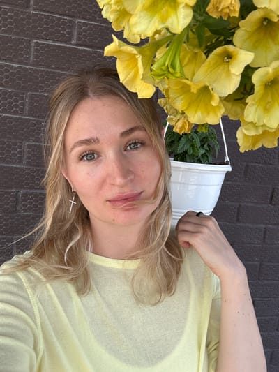 A young woman with blonde hair and green eyes poses for a selfie in front of a dark brown brick wall. She is wearing a pale yellow crewneck shirt and a silver cross earring with small rhinestones. Her right hand is resting on her shoulder, and she is wearing a black ring on her right index finger. A white pot of yellow flowers hangs in the background to her right. The scene appears to be outdoors.
