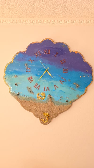 A wall clock with a cloud-like shape and a sea-themed design hangs on a textured, light orange wall. The clock face features Roman numerals in red and gold, representing the hours. The design transitions from a dark blue at the top, representing the sky, to various shades of blue and turquoise, depicting the ocean. The bottom portion of the clock is adorned with sand, seashells, and small pebbles, mimicking a beach. A golden seahorse figurine is placed on the sand at the bottom center of the clock. The clock has yellow hour and minute hands, with the time appearing to be approximately 10:50. The image was taken in the city of Crailsheim, Germany.