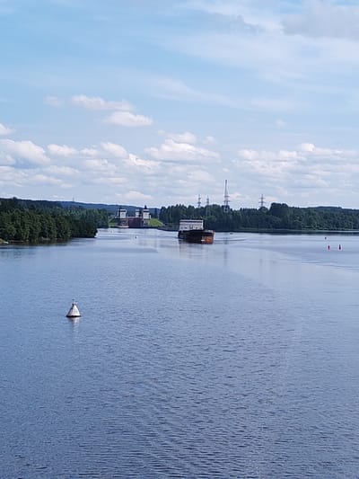 A wide, calm river flows under a partly cloudy blue sky in this daytime, landscape view. On the left side of the river, a green, tree-lined bank is visible, with a large, industrial-looking structure, possibly a lock or dam, situated on the bank further in the distance. A solitary white buoy with the number "13" is floating in the foreground of the river, and a ship is seen in the middle ground, moving away from the viewer. The ship appears to be a barge with a cabin structure on its rear. In the background, beyond the river and the trees, power lines and tall transmission towers can be seen. The overall scene suggests an industrial or navigational waterway, potentially located in or near Анхимово, Russia, given the presence of such infrastructure.