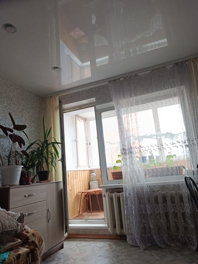 The image depicts an indoor scene, likely a living room or apartment, with a view of a balcony through a doorway. The room features a light-colored, possibly textured wallpaper and a reflective, white stretch ceiling with recessed lighting fixtures visible. A wooden cabinet with drawers and a handle stands against the left wall, partially covered by a patterned blanket and pillows. Several potted plants are placed on top of the cabinet and on a shelf near the doorway.

The doorway leads to a balcony with light-colored wooden paneling on the walls. A small, orange stool with a patterned seat sits on the balcony, and a large plastic water bottle is visible on a shelf. The balcony is framed by a window with translucent, white lace curtains, patterned with floral motifs. Through the curtains, more potted plants are visible on a windowsill. A white radiator is situated beneath the window. The floor in the room appears to be tiled with a grey and white pattern. The overall impression is of a modest, lived-in interior. The location in Gusinoozyorsk, Russia is a reasonable assumption given the details, although not definitively confirmed by the image alone.