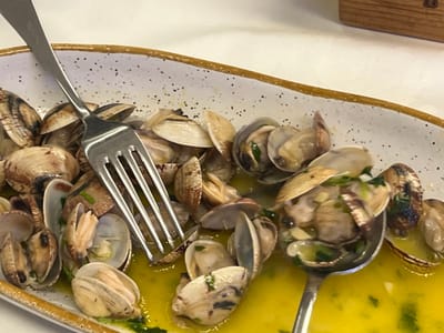 An overhead, close-up view depicts a white, oval-shaped ceramic platter filled with numerous bivalve mollusks, identified as clams, prepared and served in their shells. The clams are submerged in a vibrant yellow, opaque liquid sauce, indicative of olive oil, with visible chopped green herbs (consistent with parsley) and sliced garlic cloves. Many clam shells are open or partially open. A four-tine silver-colored metal dining fork is positioned diagonally across the left portion of the platter, resting partially on the clams and partially in the sauce. A silver-colored metal dining spoon is positioned in the right portion, partially submerged in the sauce, scooping several clams and a portion of the liquid. The platter features an irregular brown/golden rim and a white interior surface with numerous small, dark speckles. It rests on an undifferentiated white surface, likely a tablecloth or tabletop. A light-colored, vertically oriented wooden object with a darker top edge is partially visible in the upper right background. The scene takes place in a city in Caminha, Portugal.