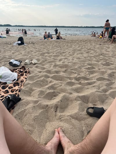 The image depicts a sunny day at a crowded beach, likely in Загародскі сельскі Савет, Belarus. The scene is dominated by sand, with the water of a lake or river in the background, where many people are swimming and enjoying the water. On the sand, several individuals are scattered, some sitting, some lying down, and others walking. There are various objects present, including towels, beach bags, and footwear, suggesting a day of leisure and recreation. The weather appears to be warm and pleasant, with clear skies and the sun casting shadows on the sand, indicating it is daytime. The overall atmosphere is one of a lively and active beach environment on a summer day.