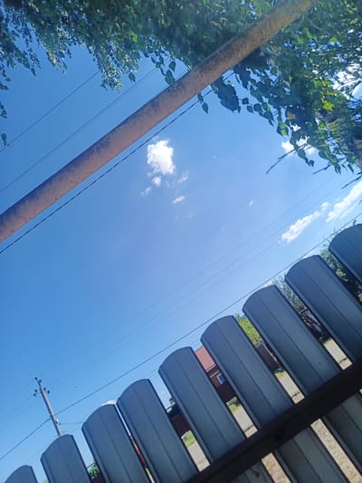The image depicts a skyward view from a fenced area in Staronizhestebliyevskaya, Russia. The foreground is dominated by a grey, corrugated metal fence, with its arched tops forming a repeating pattern that extends diagonally across the lower portion of the frame. Beyond the fence, glimpses of a residential area are visible, including a red-tiled roof and a window of a house, along with scattered greenery and pathways. Above the fence, a bright blue sky stretches out, punctuated by a few scattered white clouds. A light-colored pipe runs diagonally across the upper left of the image, parallel to the fence. Green leaves from an overhanging tree are visible in the upper part of the frame, casting shadows. Electrical wires and a utility pole are present in the middle ground, to the left of the frame. The overall impression is of an outdoor scene on a clear day, likely in a suburban or rural setting.