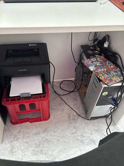 Underneath a white desk, a Samsung ML-1640 printer is positioned on a red plastic crate. A stack of white paper is visible in the printer's output tray. To the right of the printer, a computer tower is lying on its side. The tower has various stickers on its surface, including one with cartoonish characters. Power cords and other cables are strewn around the computer and printer. The scene appears to be set indoors, on a floor with a marble-like pattern. The overall impression is one of a cluttered workspace. The location is identified as Vileyka, Belarus.