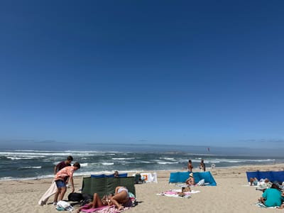 This image depicts a wide view of a sandy beach on a clear, sunny day, identified as a location in Caminha, Portugal, with the Forte da Ínsua visible in the distant ocean. The sky is a deep, cloudless blue. The ocean features moderate waves with visible whitecaps.
In the foreground, a group of approximately ten individuals is engaged in various beach activities. On the left, two young men stand near a green fabric windbreak and a dark bag. One male wears a patterned short-sleeved shirt and shorts, while the other wears a dark t-shirt and light shorts, leaning against a white and beige striped towel. Adjacent to them, two young women lie on colored towels on the sand; one is on her back, the other on her stomach, both appearing to interact with mobile phones.
Further to the right, a person lies beneath a blue fabric windbreak. In the mid-ground, near the water's edge, two men stand, one shirtless, the other in a light shirt, with a red warning flag visible behind them. Another individual is partially obscured by a windbreak in the mid-distance. On the far right foreground, a person is situated under another blue windbreak, and another individual sits upright on a towel wearing a green top.
The sandy beach extends into the distance, with faint land masses visible on the horizon to the right, beyond the choppy ocean.