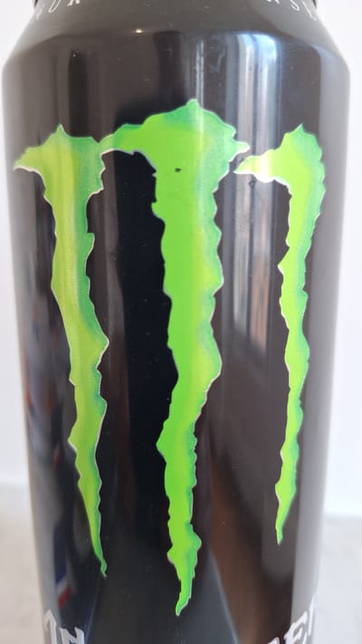 A close-up, eye-level, studio shot shows the side of a black Monster Energy drink can. The iconic green claw mark logo is prominently displayed, with three distinct "claws" of varying lengths and widths. The background is a plain, light color, likely a wall. There are no people or other objects visible in the frame. The lighting appears to be artificial, creating some reflections on the can's surface. The location is not specified as being in Fontainebleau, France, as this information cannot be determined from the image alone.